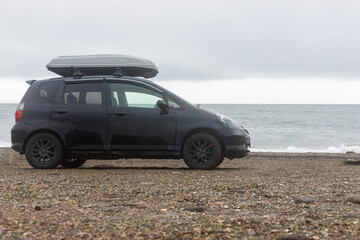 Fototapeta na wymiar A black car on the beach in summer.Car parking on the sea beach with a trunk.Car roof with luggage box On rooftop on the rack system.Summer trip or auto travel adventure concept.Copy space