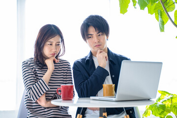 明るいリビングでパソコンを見つめ悩む若い夫婦