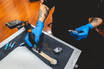 Hands of a tattoo artist holding a tattoo machine charging black ink, and work table with needles,...