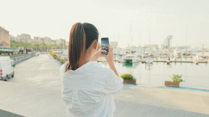 Fototapeta na wymiar Young woman taking photo of the sea port on mobile phone. Back view