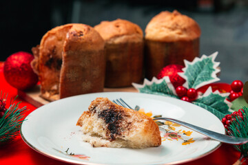 Slice of panettone on a white plate with three panettoni over a red table with various christmas decorations