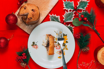 Top view of a slice of panettone on a white plate over a red table with various christmas...