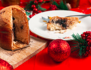 Slice of panettone on a white plate over a red table with various christmas decorations and sliced...