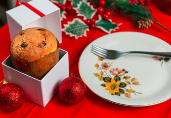 Front view of a panettone on a gift box and a white plate over a red table and various christmas...