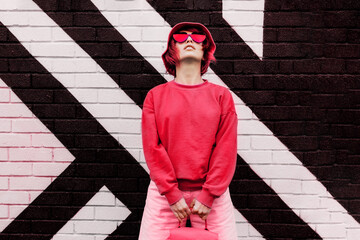 Hipster fashion young woman in trendy magenta color sweatshirt and sunglasses and bucket hat posing on the painted brick wall background. Color of the 2023 year. Urban city street fashion. - obrazy, fototapety, plakaty