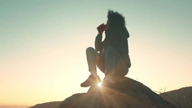 Hiker woman sits on edge of cliff against background of sunrise, drinking coffee, energy drink