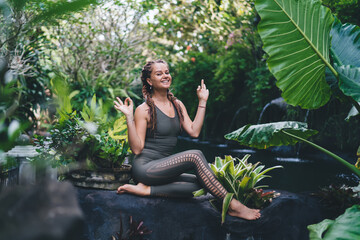 Smiling young woman sitting with raised hands and meditating in park