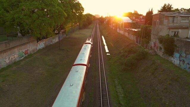 A drone flies over a train that goes through the city, in the middle of sunset

