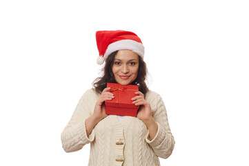 Attractive woman in Santa hat and beige sweater, holding gift box in red packaging with happy present, isolated on white background. Expectation of presents for Christmas and new year. Copy ad space