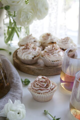 Muffins with cream and chocolate sprinkles on a wooden stand near white ranunculus and a cup of tea