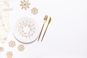Festive Christmas table setting with white plate with ornament,  golden cutlery set and decorative snow flakes on white background with copy space for your design.