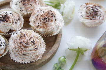 Muffins with cream and chocolate sprinkles on a wooden stand near white ranunculus and a cup of tea