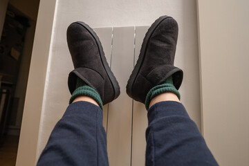 Person in warm home shoes and socks warming up near the radiator at home, energy crisis in Europe