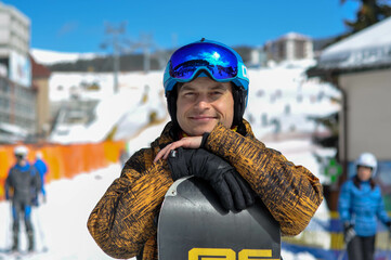 A happy and contented snowboarder stands with a snowboard near the ski lifts. Ski resort in the...