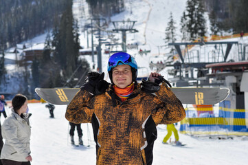 A happy and contented snowboarder stands with a snowboard near the ski lifts. Ski resort in the...