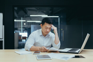 A hard day's work. A serious young Asian man sits tiredly in the office at the desk, works with documents, problems with accounts, holds his head with his hand. - obrazy, fototapety, plakaty