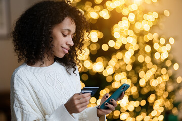 Merry Christmas and Happy Holidays! Young african american woman using smartphone for shopping online with credit card on by the Christmas tree         