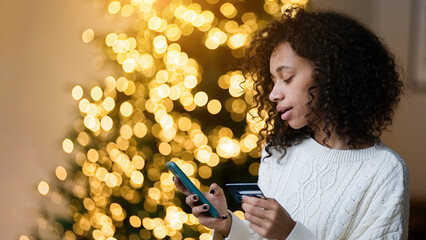 Merry Christmas and Happy Holidays! Young african american woman using smartphone for shopping online with credit card on by the Christmas tree         