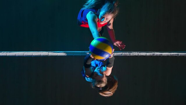 Two young female athletes playing volleyball on the playground in blue light. Top view of woman serving ball and an opponent hitting the ball and rejoicing in victory.