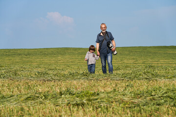 Fototapeta na wymiar Grandfather and granddaughter spending time together. An adult man and a little girl holding hands walk across the field. The man has a camera. They are happy to be together.
