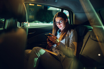 Business woman using smartphone while sitting in a backseat of a car at night - obrazy, fototapety, plakaty