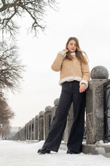 Brunette young woman in black jeans and beige sheepskin coat stands pensively in winter park. Winter look for every day