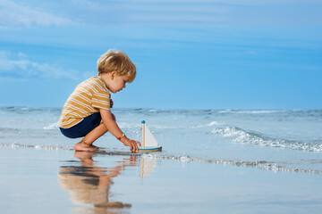 Handsome little blond boy put toy boat in the ocean waves water