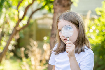 Curious clever elementary school age child, girl looking at the camera through a magnifying glass, holding a loupe in hand, one eye closed, one visible, copy space, blurred background. Young explorer