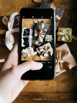 Female hand taking photo with smartphone of stylish wrapped Christmas gifts  on wooden background. The girl taking a picture of a gifts on the phone. Christmas, New Year, social media, holiday concept