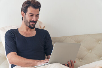 Handsome young man working on laptop while sitting on the bed in the morning at home. Freelance guy success using notebook searching about work online or shopping online. Lifestyle technology concept.