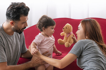 Cheerful asian mother holding teddy bear playful infants daughter with father sitting together side sofa at home. Parent use teddy bear innocence toddler laughing togetherness. Domestic life family.