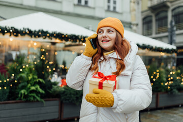 Happy young beautiful excited woman holding gift box talking on mobile phone on winter street. In...