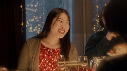 Asian woman drinks wine, talks and smile. Multi cultural friends celebrating Christmas or New Year 2023. Served holiday table. Warm atmosphere of Christmas dinner at home. Christmas party.