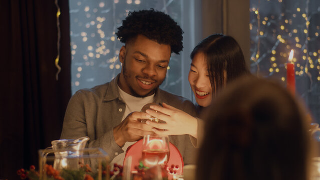 Surprised African American Man Opens Gift. Multi Cultural Friends Celebrating Christmas Or New Year 2023. Served Holiday Table With Different Dishes. Warm Atmosphere Of Christmas Dinner At Home.