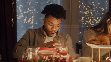 Surprised African American man opens gift. Multi cultural friends celebrating Christmas or New Year 2023. Served holiday table with different dishes. Warm atmosphere of Christmas dinner at home.