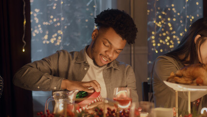 Surprised African American man opens gift. Multi cultural friends celebrating Christmas or New Year 2023. Served holiday table with different dishes. Warm atmosphere of Christmas dinner at home.