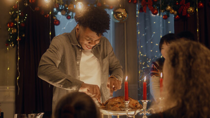 African American man cuts turkey or chicken with knife. Multi cultural friends celebrating Christmas or New Year. Served holiday table with candles. Warm atmosphere of Christmas dinner at home.