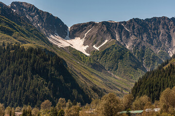 Seward, Alaska, USA - July 22, 2011: Landscape under blue sky of dark mountain range with snow patches in crevices and green forest on lower flanks. Town roofs at bottom