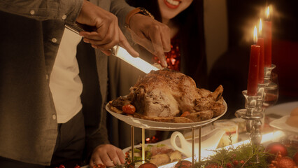 Close-up shot of African American man cutting turkey or chicken. Multi cultural family celebrating Christmas or Thanksgiving Day. Served table with dishes and candles. Family Christmas dinner at home.
