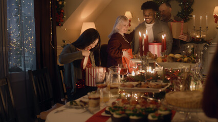 Happy multi cultural family celebrating Christmas or New Year 2023, exchanging gifts. Served holiday table with different dishes and candles. Warm atmosphere of family Christmas dinner at home.