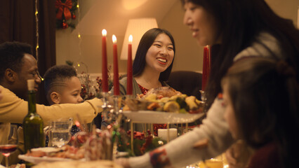 Happy multi cultural family celebrating Christmas or New Year 2023, talking and eating. Served holiday table with different dishes and candles. Warm atmosphere of family Christmas dinner at home.