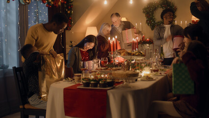 Happy multi cultural family celebrating Christmas or New Year 2023, exchanging gifts. Served holiday table with different dishes and candles. Warm atmosphere of family Christmas dinner at home.