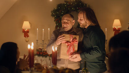 Man giving gift to his senior father. Happy family celebrating Christmas or New Year 2023. Served holiday table with candles. Warm atmosphere of family Christmas dinner at home.