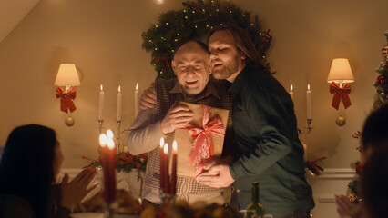 Fototapeta na wymiar Man giving gift to his senior father. Happy family celebrating Christmas or New Year 2023. Served holiday table with candles. Warm atmosphere of family Christmas dinner at home.