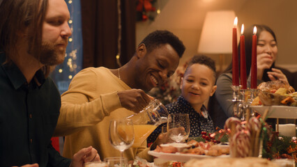 Happy multi cultural family celebrating Christmas or New Year 2023. African American young boy laughs. Served holiday table with dishes and candles. Warm atmosphere of family Christmas dinner at home.