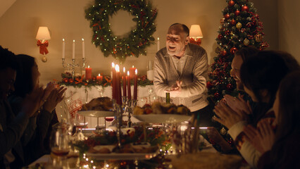 Elderly grandfather tells joke or life story to his large multi cultural family. They celebrating Christmas. Served table with dishes and candles. Warm atmosphere of family Christmas dinner at home.