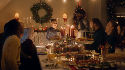 Fototapeta na wymiar African American man givs gift to young boy. Happy family celebrating Christmas or New Year 2023. Served holiday table with dishes and candles. Warm atmosphere of family Christmas dinner at home.