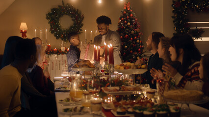 African American man givs gift to young boy. Happy family celebrating Christmas or New Year 2023. Served holiday table with dishes and candles. Warm atmosphere of family Christmas dinner at home.