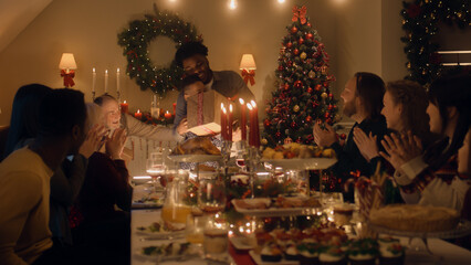 African American man givs gift to young boy. Happy family celebrating Christmas or New Year 2023. Served holiday table with dishes and candles. Warm atmosphere of family Christmas dinner at home.