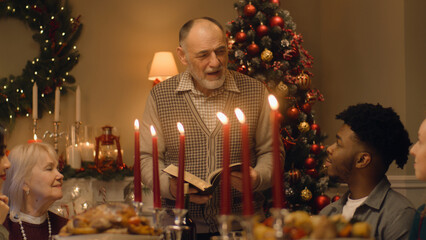 Obraz na płótnie Canvas Drandfather reads Bible on family Christmas dinner. Large diverse family praying before celebrating Christmas or New Year 2023. Served holiday table with delicious meal and candles. Winter holidays.